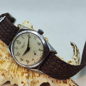 May include: A vintage wristwatch with a silver case and a cream face, displaying black numerals and hands. It has a brown woven strap. The watch is presented on a seashell.