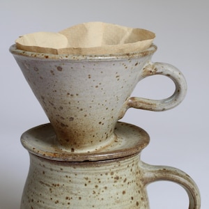 Könnte beinhalten: Ein gesprenkelter Keramik-Kaffeefilterhalter mit Henkel, der auf einer passenden gesprenkelten Keramiktasse steht. Der Filterhalter enthält einen hellbraunen Papierkaffeefilter. Die Keramik hat eine hellbeige Glasur mit braunen Sprenkeln.