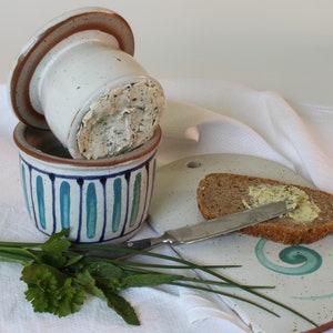 Könnte beinhalten: Eine weiße Keramik-Butterdose mit blau-grünem Streifenmuster. Die Dose ist mit einer cremigen Buttermasse gefüllt. Eine Scheibe Brot mit Butter bestrichen liegt auf einem weißen Keramik-Schneidebrett mit einem blau-grünen Wirbelmuster. Ein Bund grüner Schnittlauch und ein Buttermesser liegen ebenfalls auf dem Tisch.