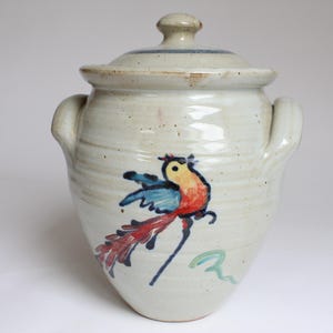 Könnte beinhalten: Keramiktopf mit Deckel, mit handgemaltem Vogelmotiv in Rot, Blau und Gelb. Der Topf hat zwei Griffe und eine gesprenkelte, cremefarbene Oberfläche. Der Deckel hat einen blauen Streifen.