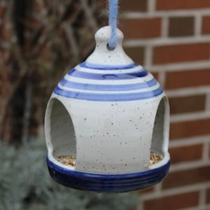 Könnte beinhalten: Ein Vogelhäuschen aus Keramik mit gesprenkelter cremeweißer Glasur und blauen horizontalen Streifen. Das kuppelförmige Futterhaus hat zwei große bogenförmige Öffnungen und einen soliden blauen Boden. Ein hellblauer Wollfaden ist oben durchgezogen.