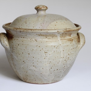Könnte beinhalten: Ein gesprenkelter, cremefarbener Keramiktopf mit Deckel und zwei seitlichen Griffen. Der Deckel hat einen kleinen Knopf. Der Topf hat ein rustikales, handgemachtes Aussehen mit braunen Sprenkeln und einer leicht unebenen Oberfläche.