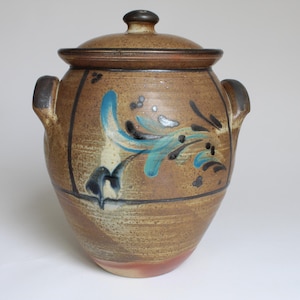 Könnte beinhalten: Ein Keramiktopf mit Deckel, mit einer braunen und beigefarbenen gesprenkelten Oberfläche. Der Topf ist mit blauen und schwarzen floralen Designs verziert. Er hat zwei seitliche Griffe und einen dunkelbraunen Knopf auf dem Deckel.