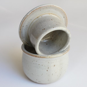 Könnte beinhalten: Ein Keramik-Butterdose mit Deckel. Die Dose und der Deckel sind in einem gesprenkelten, cremefarbenen Farbton gehalten und haben eine leicht raue Textur. Der Deckel liegt verkehrt herum auf der Dose. Die Dose ist rund.