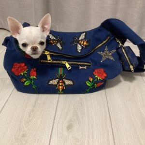 Könnte beinhalten: Ein weißer Chihuahua-Hund schaut aus einer blauen Denim-Haustiertasche. Die Tasche ist mit gestickten Bienen, Rosen und einem Stern verziert, mit goldenen Reißverschlüssen und einer kleinen passenden Tasche.