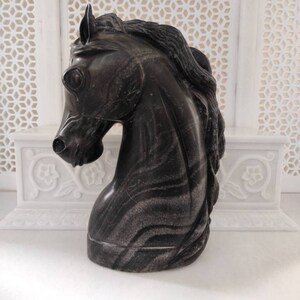 May include: A black marble horse head sculpture with a detailed mane. The sculpture is on a white marble surface.