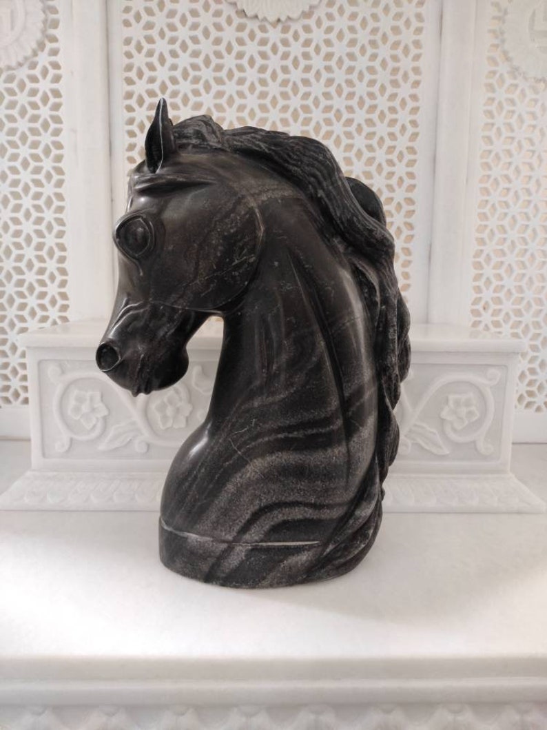 May include: A black marble horse head sculpture with a detailed mane. The sculpture is on a white marble surface.