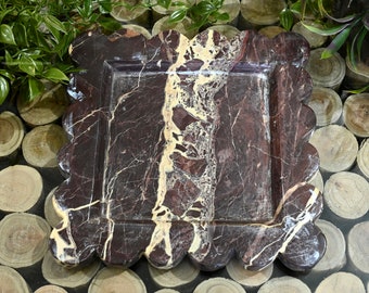 May include: A square, scalloped-edge serving tray made of dark brown marble with white and cream colored veins. The tray is sitting on a surface of stacked wooden rounds.