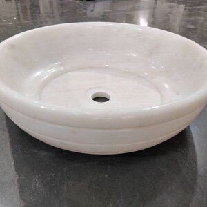 May include: A white marble bathroom sink with a round bowl and a single drain hole. The sink has a smooth, polished finish and a slightly raised rim.