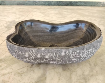 May include: A black stone sink with a rough, textured exterior and a smooth, polished interior. The sink is oval-shaped and has a unique, organic design.