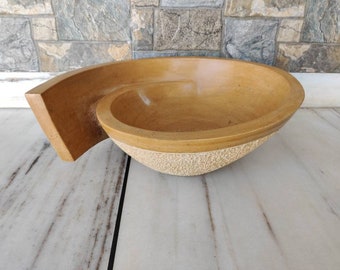 May include: A handcrafted, light brown stone bowl with a unique spiral design. The bowl has a smooth interior and a textured exterior base. The bowl's dimensions are approximately 15 inches long, 10 inches wide, and 4 inches deep.
