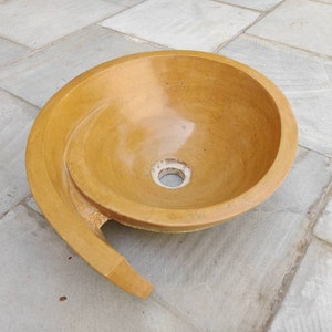 K&ouml;nnte beinhalten: Ein gelb get&ouml;ntes Steinwaschbecken mit einem Spiraldesign. Das Waschbecken hat ein kreisf&ouml;rmiges Becken und einen gebogenen, verl&auml;ngerten Rand. Das Abflussloch ist in der Mitte sichtbar. Das Waschbecken besteht aus einem festen, nat&uuml;rlichen Material.