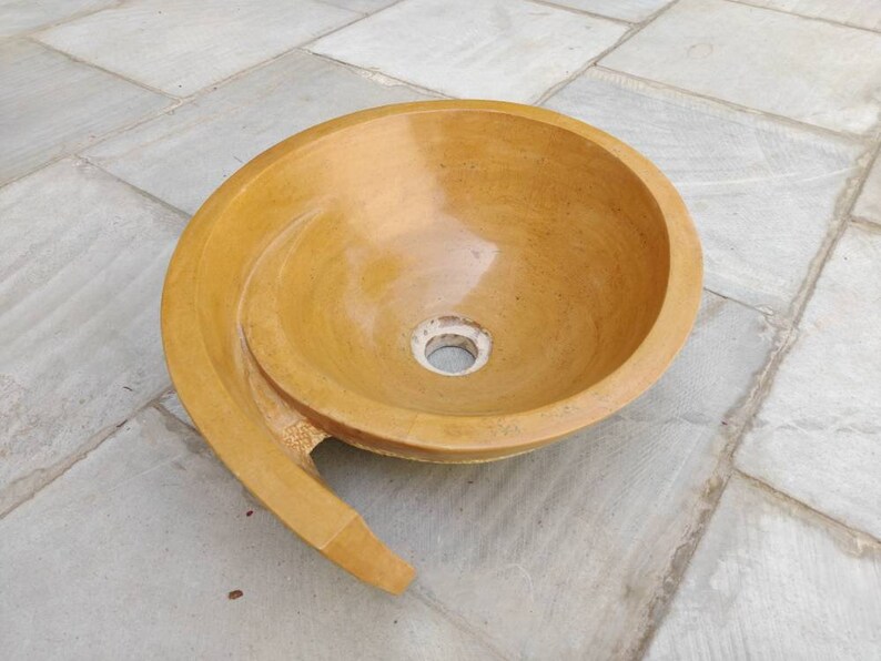 K&ouml;nnte beinhalten: Ein gelb get&ouml;ntes Steinwaschbecken mit einem Spiraldesign. Das Waschbecken hat ein kreisf&ouml;rmiges Becken und einen gebogenen, verl&auml;ngerten Rand. Das Abflussloch ist in der Mitte sichtbar. Das Waschbecken besteht aus einem festen, nat&uuml;rlichen Material.
