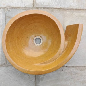 K&ouml;nnte beinhalten: Ein einzigartiges, spiralf&ouml;rmiges Keramikwaschbecken in einem warmen, erdigen Farbton. Das Waschbecken hat eine glatte, gl&auml;nzende Oberfl&auml;che und ein zentrales Abflussloch. Das Design umfasst eine gebogene, hornartige Verl&auml;ngerung. Das Waschbecken ist an einer hellgrauen gefliesten Wand befestigt.