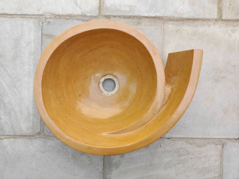 K&ouml;nnte beinhalten: Ein einzigartiges, spiralf&ouml;rmiges Keramikwaschbecken in einem warmen, erdigen Farbton. Das Waschbecken hat eine glatte, gl&auml;nzende Oberfl&auml;che und ein zentrales Abflussloch. Das Design umfasst eine gebogene, hornartige Verl&auml;ngerung. Das Waschbecken ist an einer hellgrauen gefliesten Wand befestigt.