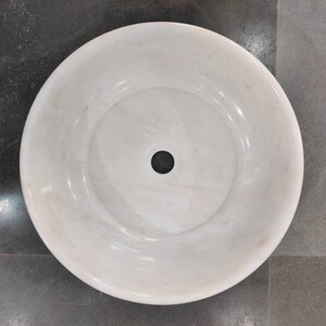 May include: A white marble sink with a round shape and a single drain hole in the center.