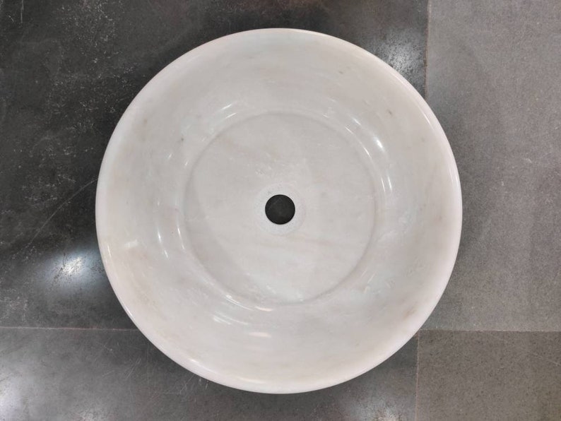May include: A white marble sink with a round shape and a single drain hole in the center.