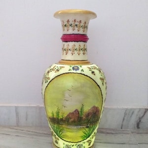 May include: A hand-painted ceramic vase with a landscape scene. The vase is decorated with gold and pink accents and features a green, brown, and blue landscape with birds flying over a lake.