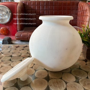 May include: White ceramic jar with a lid. The jar is round and has a smooth surface. The lid is flat and has a small knob on top. The jar is sitting on a table made of wood rounds.