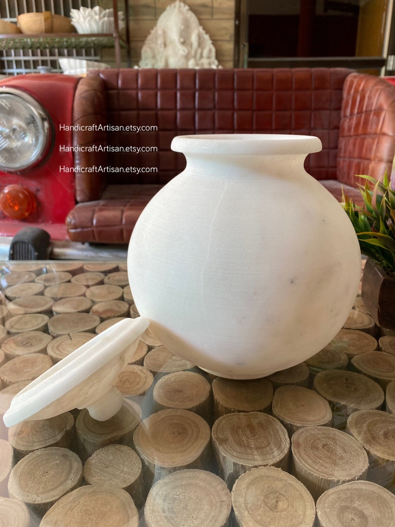 May include: White ceramic jar with a lid. The jar is round and has a smooth surface. The lid is flat and has a small knob on top. The jar is sitting on a table made of wood rounds.