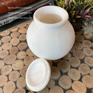 May include: White marble jar with a lid. The jar is round and has a smooth surface. The lid is also round and has a smooth surface. The jar is sitting on a table with a wooden surface.
