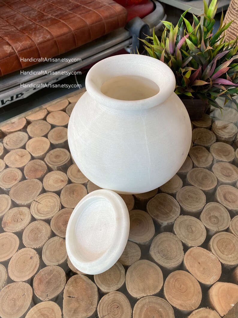 May include: White marble jar with a lid. The jar is round and has a smooth surface. The lid is also round and has a smooth surface. The jar is sitting on a table with a wooden surface.