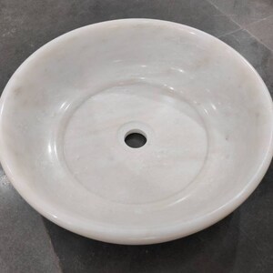 May include: A white marble sink with a round shape and a single drain hole in the center.