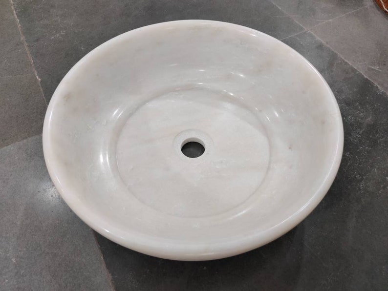 May include: A white marble sink with a round shape and a single drain hole in the center.