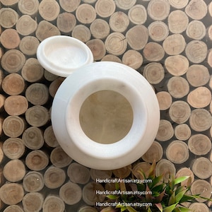 May include: White marble jar with a lid, the jar has a round opening at the top. The jar is sitting on a surface made of many small wooden circles.
