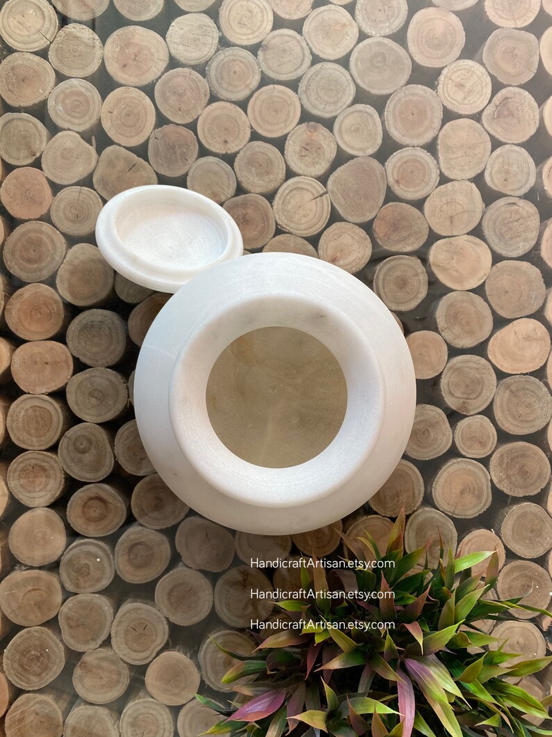 May include: White marble jar with a lid, the jar has a round opening at the top. The jar is sitting on a surface made of many small wooden circles.