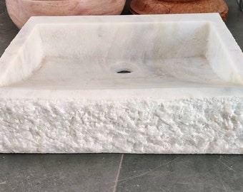 May include: A white marble sink with a rough, textured exterior. The sink has a square shape and a single drain hole in the center.