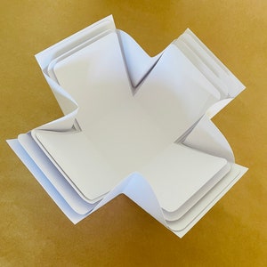 May include: A white paper box with a unique folded design, creating a star-shaped opening. The box is folded flat and ready to be assembled.