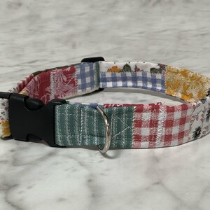 May include: A patchwork dog collar with a black plastic buckle and a silver metal D-ring. The collar features a variety of fabric patterns, including floral, gingham, and striped designs in red, blue, yellow, and green.
