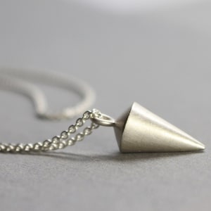 Puede incluir: Un collar con colgante plateado en forma de cono. El colgante está unido a una delicada cadena. El collar se muestra sobre un fondo gris, resaltando el diseño minimalista de la joya.