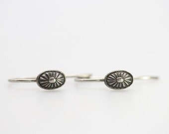 May include: A pair of silver hoop earrings with a decorative oval design. The oval is textured with a sunburst pattern and a small circle in the center.