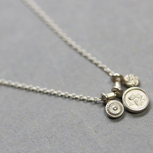 May include: A silver necklace with three charms: a small round charm with a raised design, a larger round charm with a raised image of an angel, and a small round charm with a raised design.