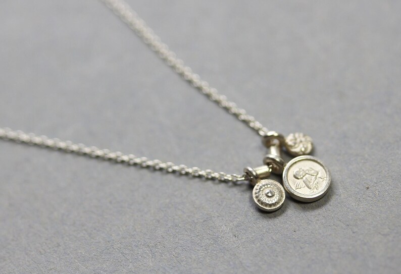 May include: A silver necklace with three charms: a small round charm with a raised design, a larger round charm with a raised image of an angel, and a small round charm with a raised design.