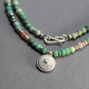 May include: A beaded necklace with turquoise and green beads, interspersed with silver-colored beads and a small pendant. The pendant is a silver-colored, circular charm with a detailed floral design. The necklace has a silver clasp.