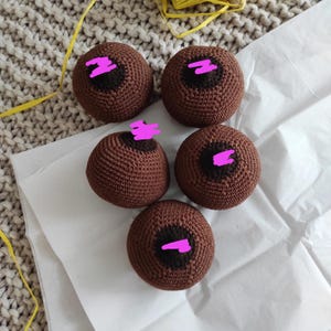 May include: Five handmade crochet spheres in varying sizes, each featuring a dark brown areola and nipple detail. The spheres are a medium brown color and rest on a white paper surface. The background includes a textured beige surface and yellow yarn.