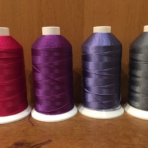 Könnte beinhalten: Vier Garnrollen in verschiedenen Farben: rosa, lila, blau und grau. Die Rollen befinden sich auf einer Holzoberfläche.