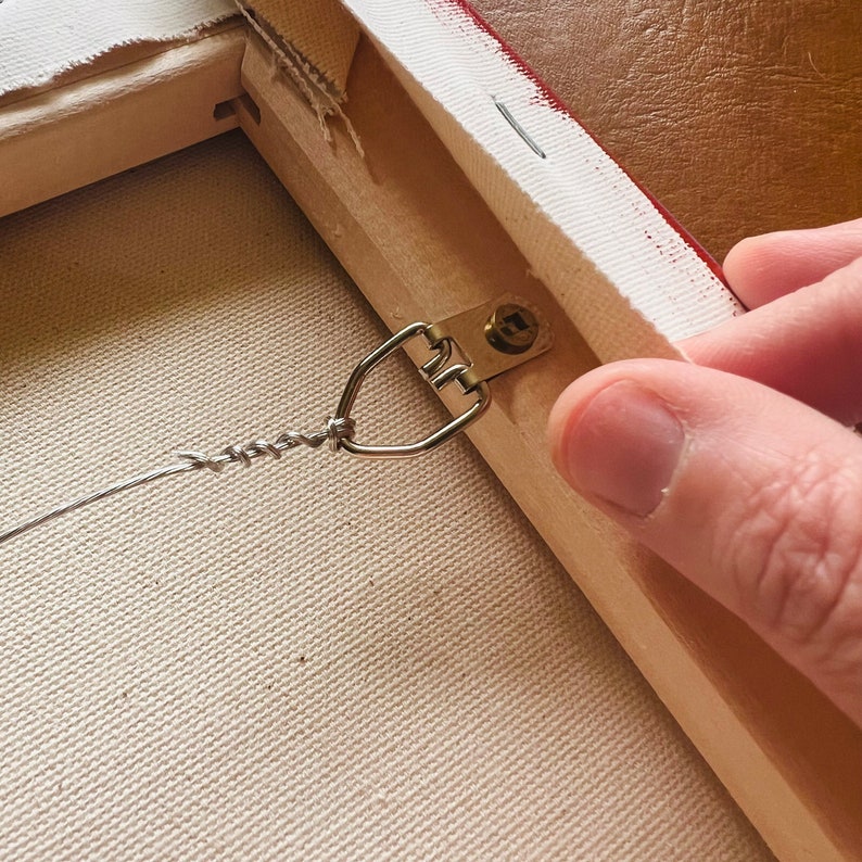 Op de afbeelding: Detail van de achterkant van een canvas kunstwerk. Een metalen D-ring hanger is met een schroef aan het houten frame bevestigd. Een gedraaide draad is aan de D-ring bevestigd, klaar om op te hangen. Het canvas is over het houten frame gespannen.