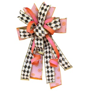 May include: A decorative bow made of wired ribbon featuring a black and white diamond harlequin pattern, solid neon orange ribbon, and a light pink ribbon with a repeating orange sunburst or floral pattern, finished with gold wire edges.
