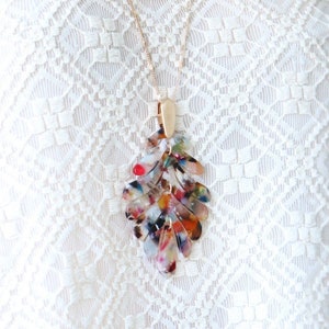 May include: A gold chain necklace with a multi-colored leaf pendant. The pendant is made of clear plastic with red, orange, yellow, green, blue, and purple accents. The necklace is worn by a person wearing a white lace top.