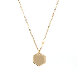 May include: A gold-colored necklace with a hexagonal pendant. The chain is delicate, with small green beads and gold-colored accents. The pendant is a smooth, polished gold color. The necklace is displayed against a white background.