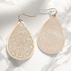 Peut inclure: Une paire de boucles d'oreilles dorées en forme de goutte d'eau. Chaque boucle d'oreille présente un motif ajouré et délicat à l'intérieur d'une forme de goutte d'eau. Le motif complexe est visible sur un fond clair, et les boucles d'oreilles ont une fermeture à crochet simple.