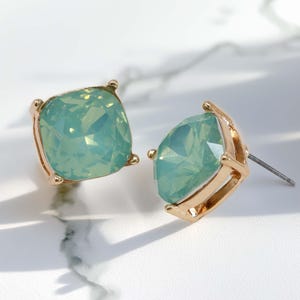 A pair of square, cushion-cut stud earrings featuring bright mint opal glass stones with fine glitter inside. The stones are set in a shiny gold-toned four-prong metal setting.
