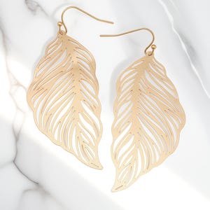 May include: A pair of gold-colored feather-shaped earrings. The earrings have a delicate, openwork design with intricate details resembling the veins of a feather. The earrings hang from gold-colored ear wires, and are displayed against a white marble background.