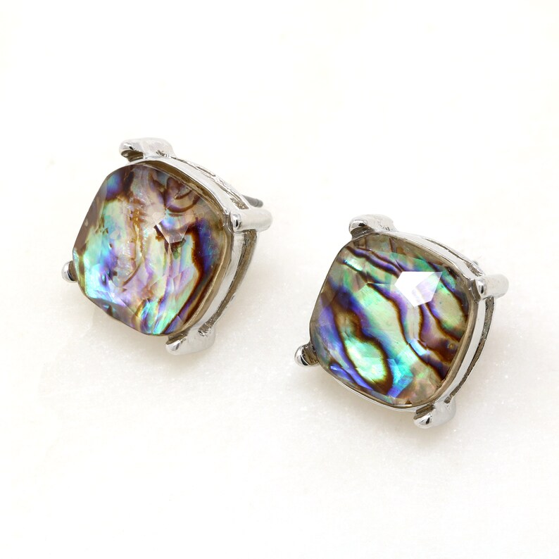 Puede incluir: Dos pendientes de bot&oacute;n cuadrados con conchas de abul&oacute;n iridiscentes engastadas en metal plateado. Las conchas de abul&oacute;n muestran una mezcla de tonos azules, verdes y morados. Los pendientes tienen un dise&ntilde;o sencillo y elegante.
