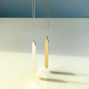 May include: Two minimalist necklaces, one silver-toned and one gold-toned, each featuring a long, slender, rectangular pendant suspended from a delicate chain. The pendants have rounded edges and hang against a soft, neutral background.
