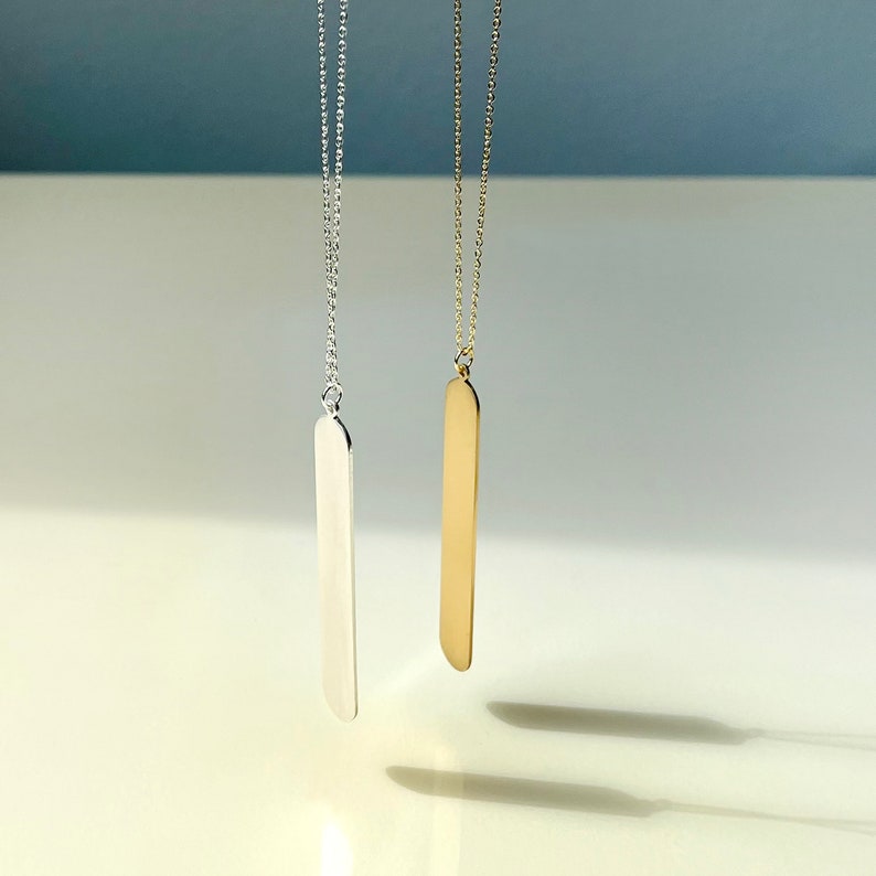 May include: Two minimalist necklaces, one silver-toned and one gold-toned, each featuring a long, slender, rectangular pendant suspended from a delicate chain. The pendants have rounded edges and hang against a soft, neutral background.